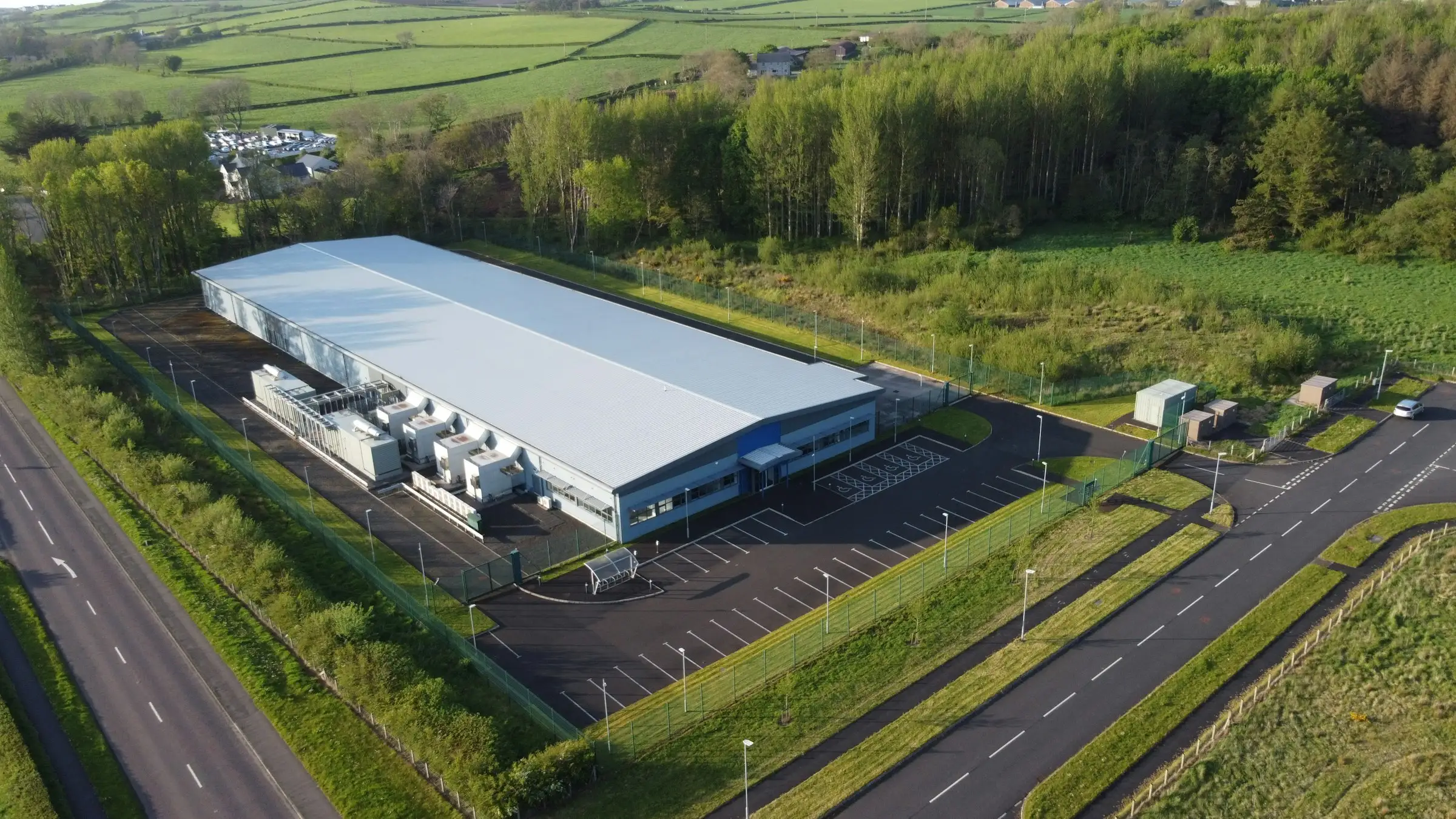 Aerial view of a data center: a long windowless warehouse ringed by security fencing, parking, and rows of cooling units, built on open green farmland.